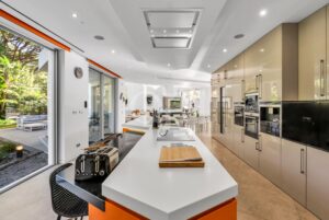 Open-plan modern kitchen with glossy beige cabinets, white island, and stainless-steel appliances; living area visible in the background through glass doors.