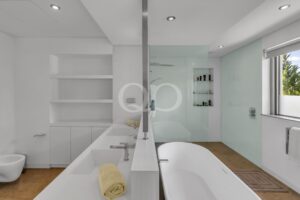 Modern white bathroom with a double vanity, large mirror, glass shower, and a window showing greenery outside.