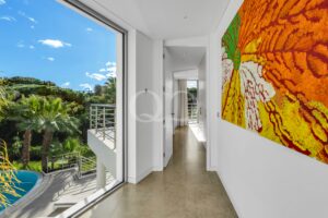 Bright corridor with white walls and a large orange-yellow mural on the right; glass doors lead to a sunny balcony with trees outside.