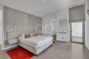 Modern hotel bedroom with a white upholstered bed, grey textured wall, and red rug by the bedside.