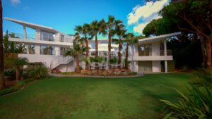 Modern white two-story house with large balconies, palm trees, and a manicured lawn in the foreground.