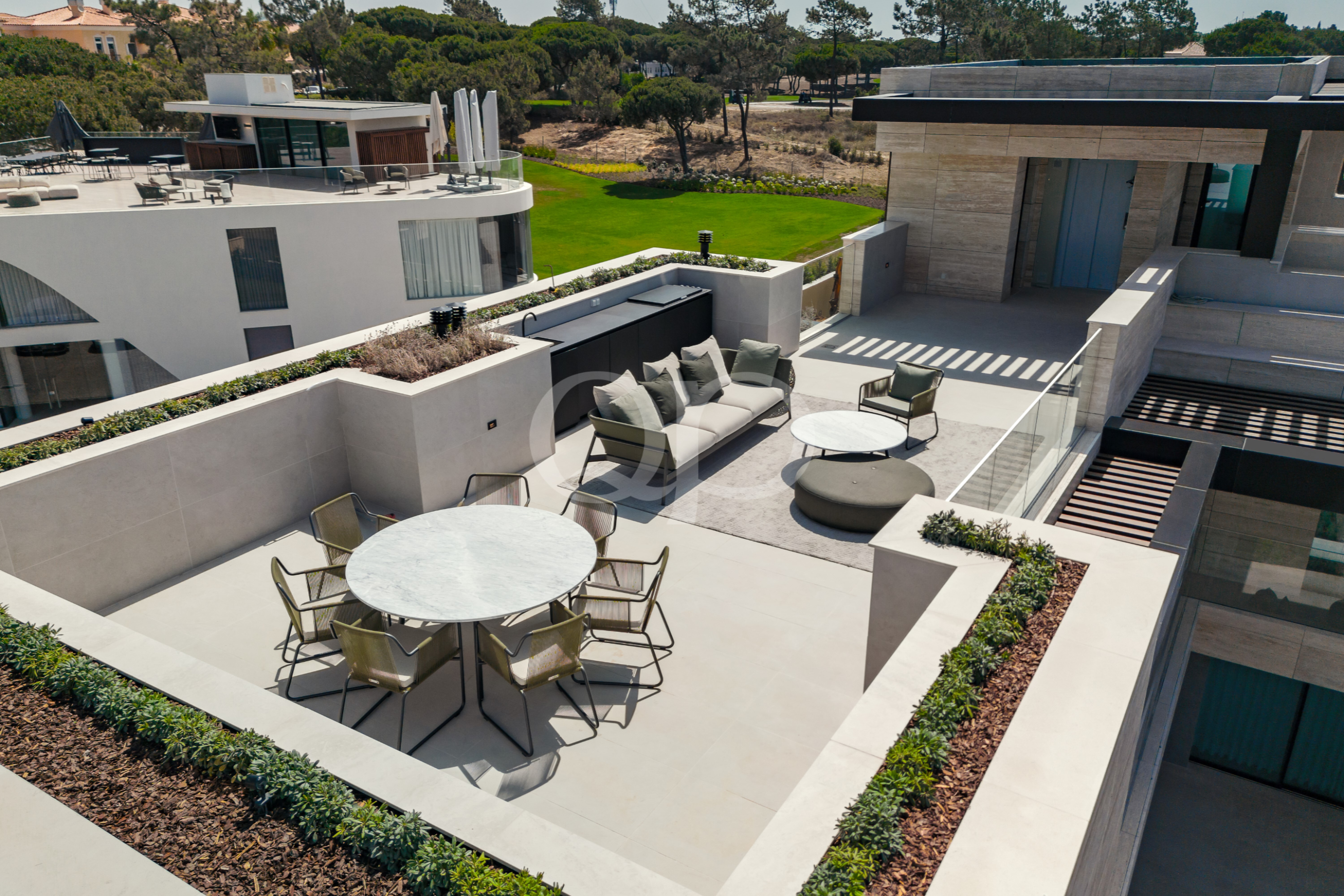 Une terrasse moderne sur le toit, agrémentée de tables rondes en marbre, de chaises beiges et d'un canapé modulable gris avec des coussins, donnant sur une pelouse verdoyante.