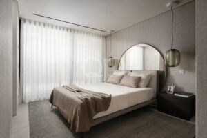 Chambre à coucher moderne de style hôtelier, avec un lit beige, des rideaux blancs vaporeux, un miroir mural rond et des suspensions en verre.