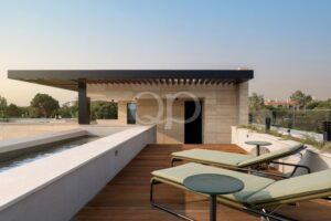 Une terrasse sur le toit avec un plancher en bois, des murs en pierre beige et une rangée de chaises longues rembourrées de couleur verte donnant sur un jardin au loin.