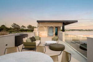 Un salon sur le toit avec des tables rondes en marbre, des chaises beiges et vert d'olive, une balustrade en verre et une pergola moderne se découpant sur le ciel au coucher du soleil.