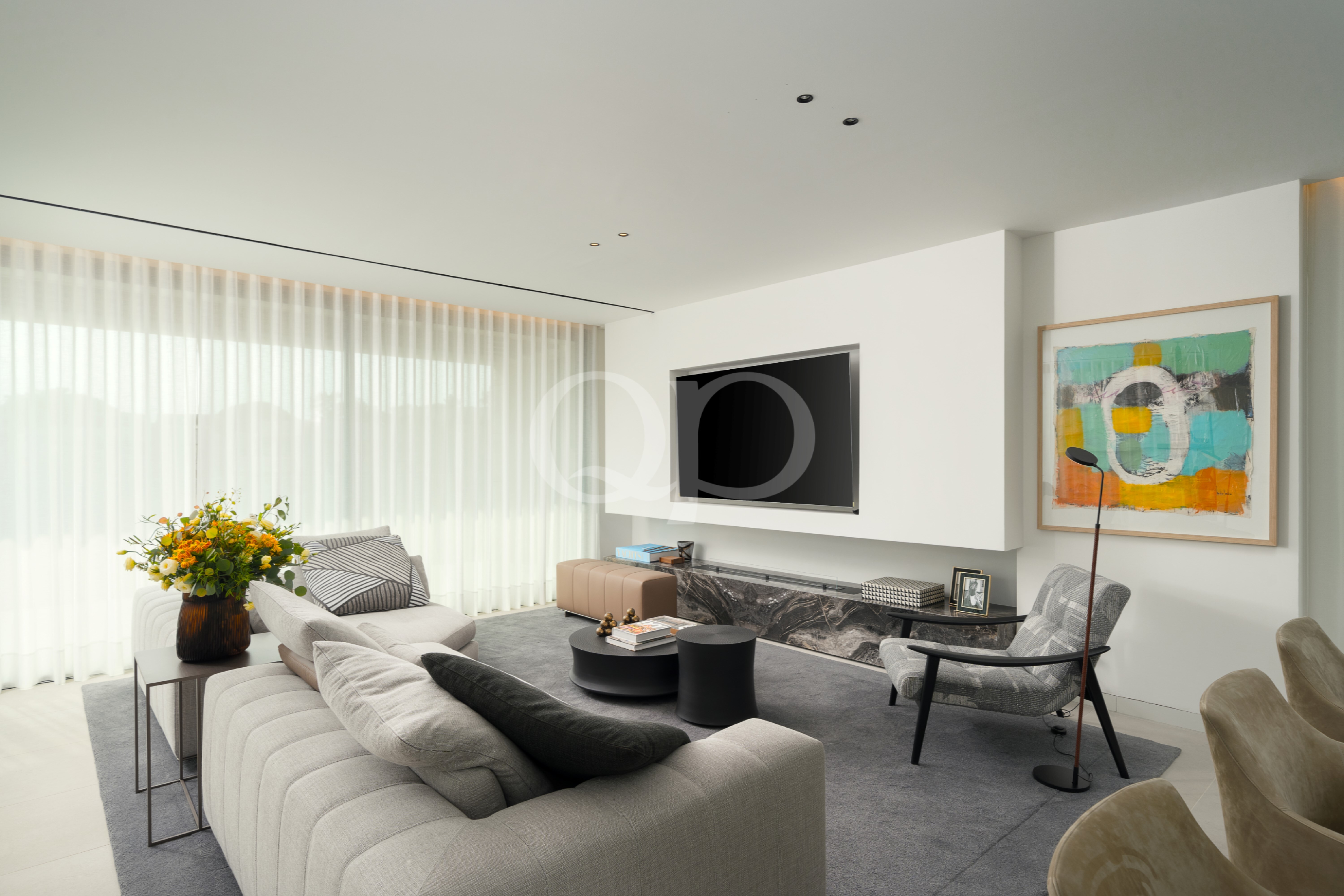 Un salon lumineux et moderne, équipé d'une télévision fixée au mur, d'un tableau abstrait aux couleurs vives et d'un canapé modulable gris clair, le tout près de grandes fenêtres ornées de voilages.