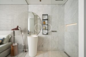 Salle de bains moderne avec un miroir rond, des murs en marbre et une étagère ouverte noire sur laquelle sont rangés des serviettes et des articles de toilette.