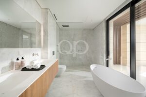 Salle de bains luxueuse en marbre, dotée d'un long plan de travail blanc, d'un meuble-lavabo en bois, de serviettes disposées sur un plateau et d'une baignoire îlot située à côté de grandes portes vitrées donnant sur une terrasse extérieure en bois.
