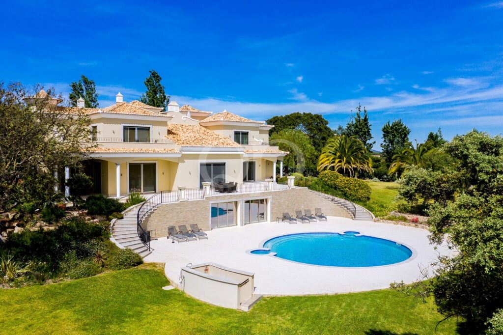 Moradia de luxo com paredes de estuque branco, telhas de terracota e uma piscina azul com formato curvo num jardim bem cuidado.