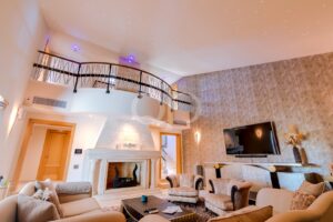 Sala de estar luxuosa com sofás bege, uma lareira de mármore e uma televisão fixada na parede, sob o corrimão curvo do mezanino.