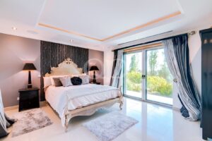 Quarto luxuoso com uma cama creme ornamentada, uma parede de destaque com padrão preto e dois candeeiros de cabeceira a condizer, junto às portas de vidro que dão para o jardim.