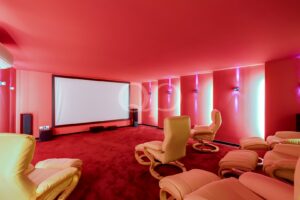 Sala de cinema em casa com tons de vermelho, dotada de um grande ecrã de projeção branco e cadeiras reclináveis em bege.