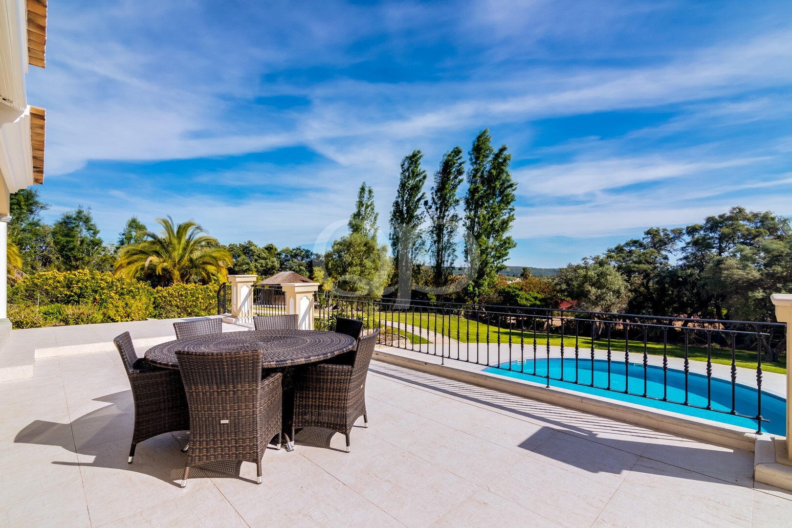 Pátio exterior com uma mesa redonda de vime e seis cadeiras, com vista para uma piscina e um jardim exuberante, sob um céu azul brilhante.