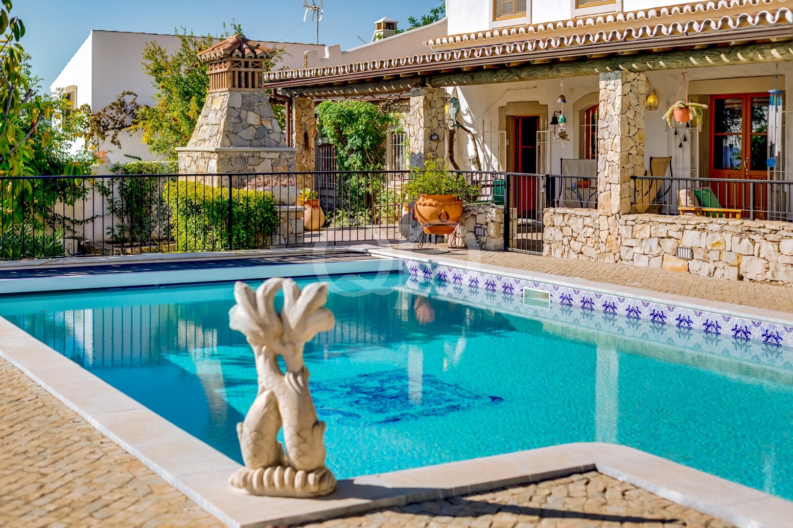 Um quintal ensolarado com uma piscina retangular em tons de turquesa, um pátio em pedra e uma villa rústica com um alpendre coberto; uma estátua de um dragão em forma de fonte encontra-se à beira da piscina.