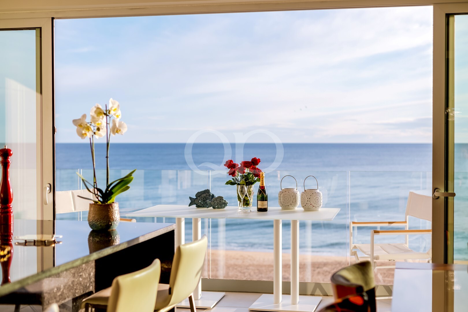 Área de refeições à beira-mar com uma mesa de vidro, orquídeas, flores vermelhas, uma garrafa de champanhe e uma vista para o oceano azul através de grandes janelas.