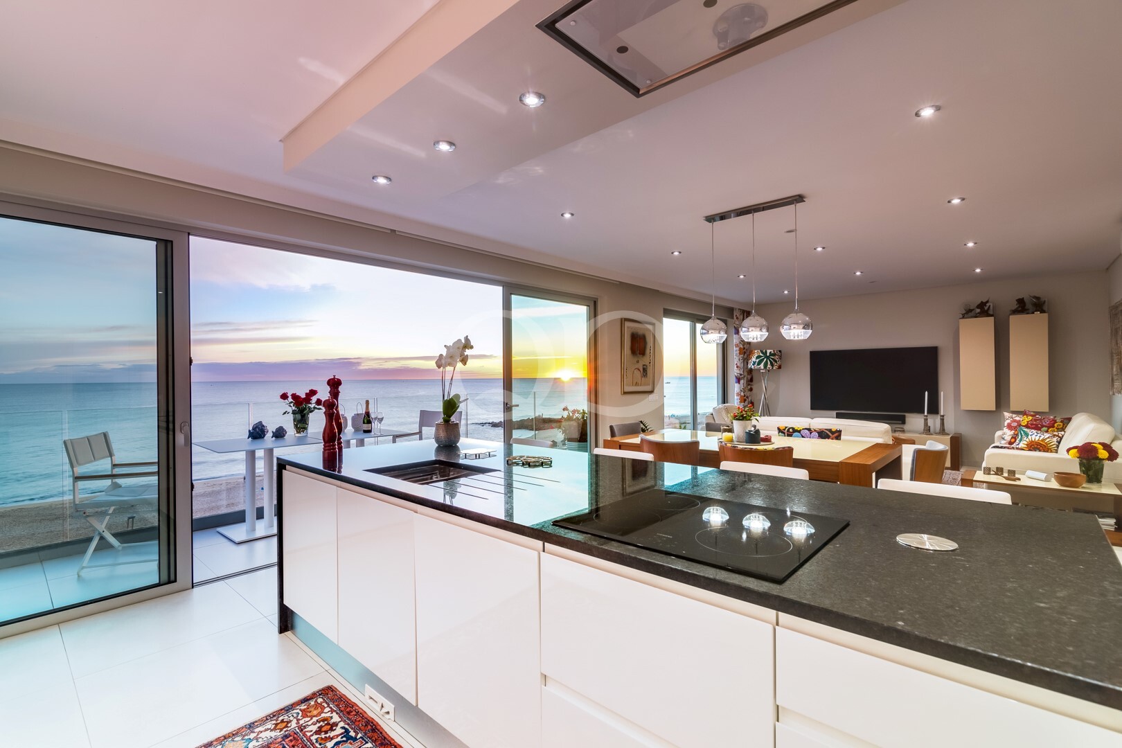 Cozinha e sala de estar em plano aberto, com armários brancos, ilha com bancada escura, placa de indução em vidro e janelas com vista para o mar ao pôr-do-sol.