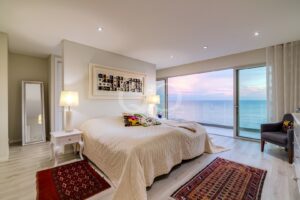 Quarto com vista para a costa, com uma cama acolchoada em tom creme, duas mesas de cabeceira com candeeiros e uma porta de vidro que dá para a varanda, com vista para o oceano ao pôr-do-sol.