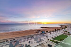 Pôr-do-sol sobre um mar calmo numa praia de areia com um passeio ladeado por palmeiras e uma fileira de guarda-sóis ao longo da costa.