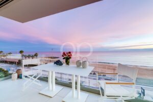 Mesa de jantar branca na varanda com cadeiras a condizer, um arranjo de rosas vermelhas no centro da mesa, com vista para um pôr-do-sol cor-de-rosa sobre a praia. (informativo)