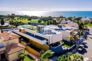 Aerial view of a modern white villa with a rooftop garden and pool overlooking the sea in a coastal neighborhood.