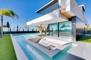Modern white, two-story home with glass balconies, a long rectangular pool, and palm trees in a landscaped yard.