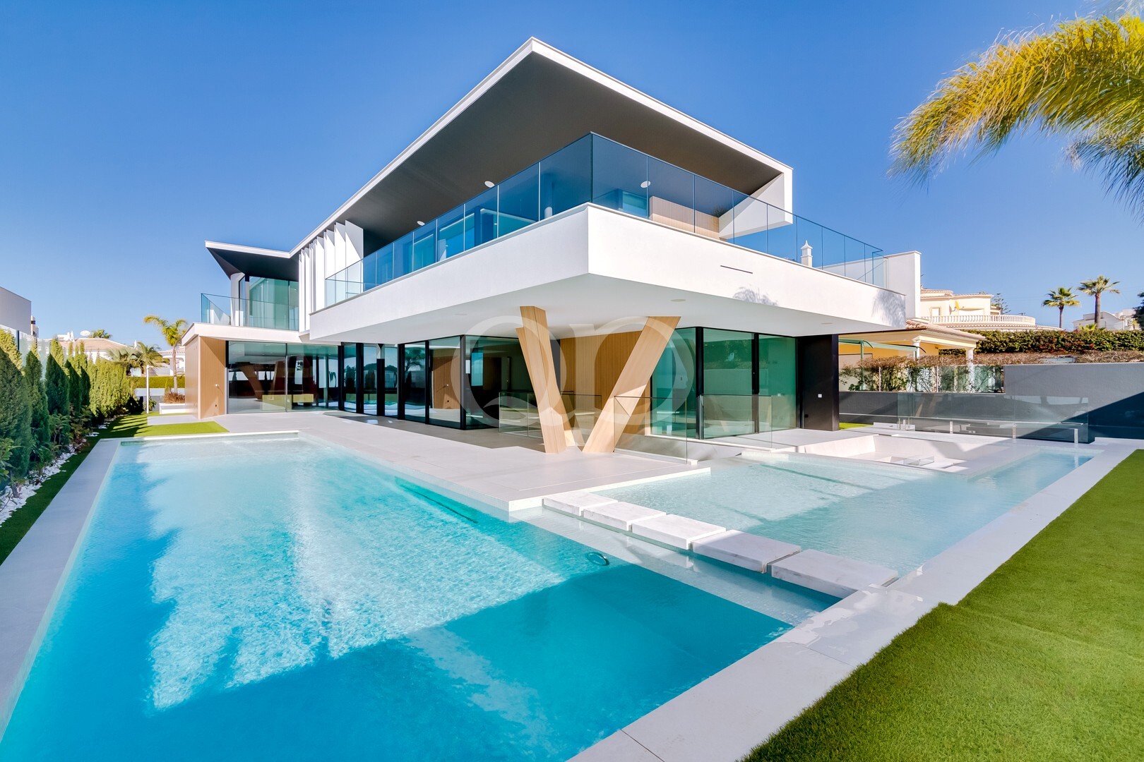 Villa moderne blanche à deux étages, avec des balustrades en verre, des poteaux en bois en forme de V et une piscine en L sous un ciel d'un bleu limpide.