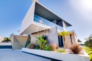 Modern two-story house with angular white and gray façades, glass balcony, and tropical landscaping under a clear blue sky.