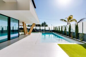 Modern backyard with a rectangular pool, white tile decking, and glass doors under a cantilevered patio.