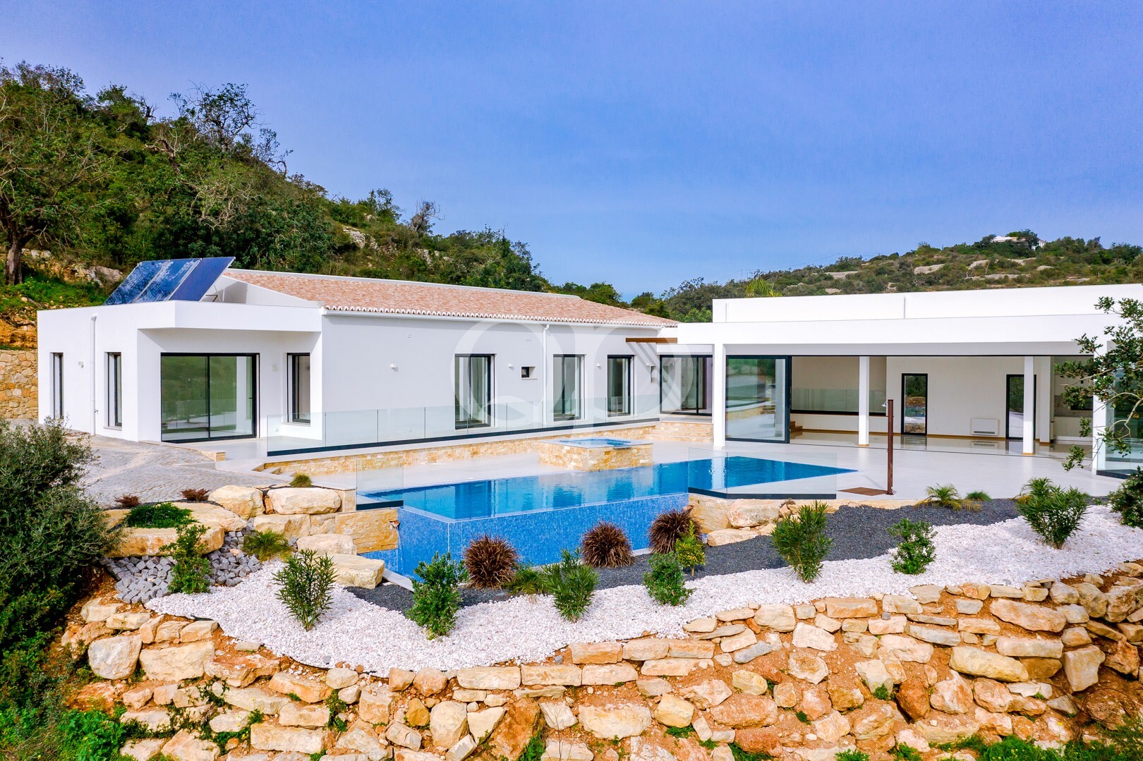 Villa moderne blanche dotée d'une balustrade en verre, d'une piscine extérieure et d'une terrasse en pierre donnant sur un jardin à flanc de colline.