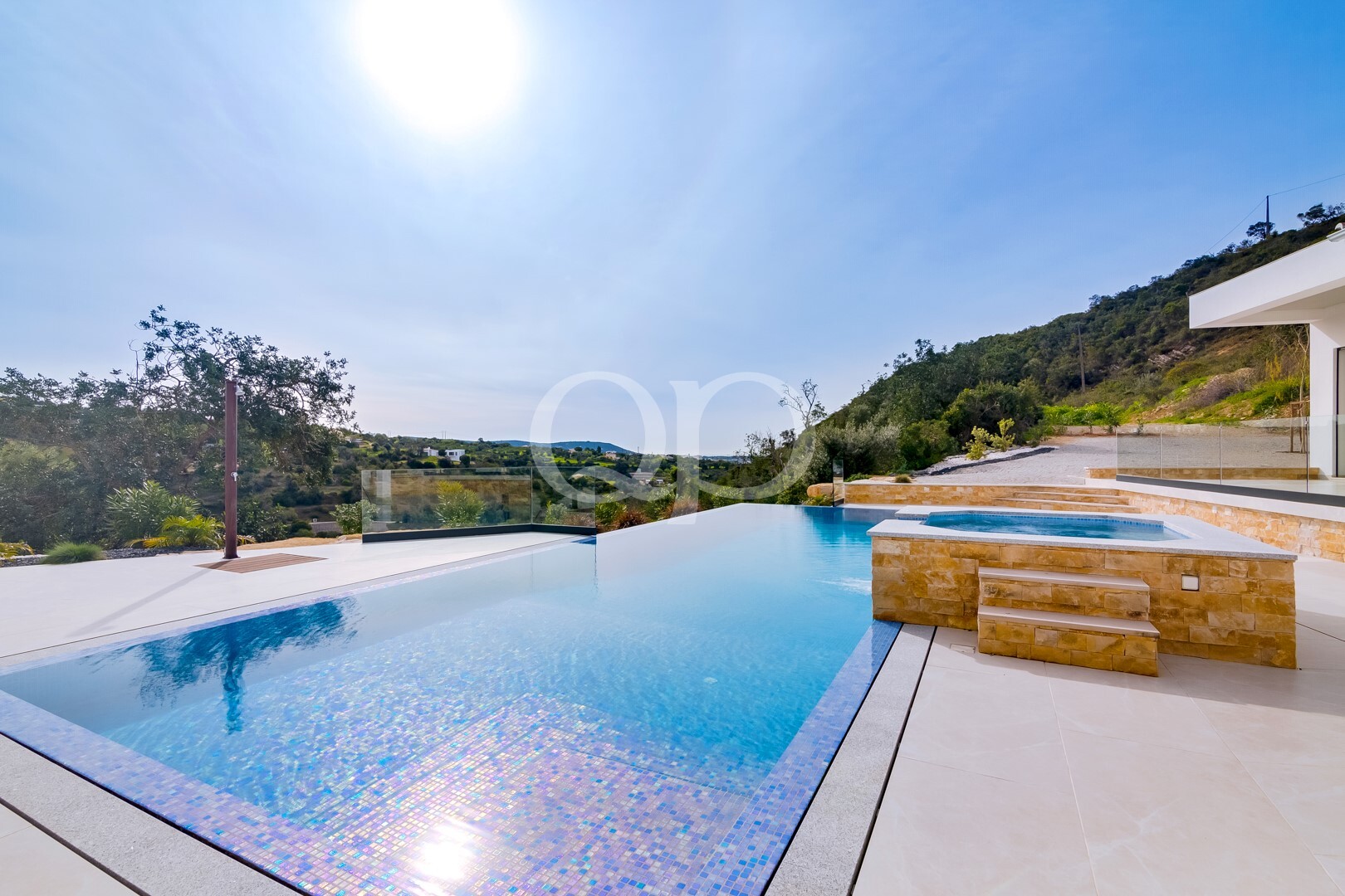 Infinity-edge pool with mosaic tiles, glass railing, and a stone-clad spa overlook a hillside landscape on a sunny day