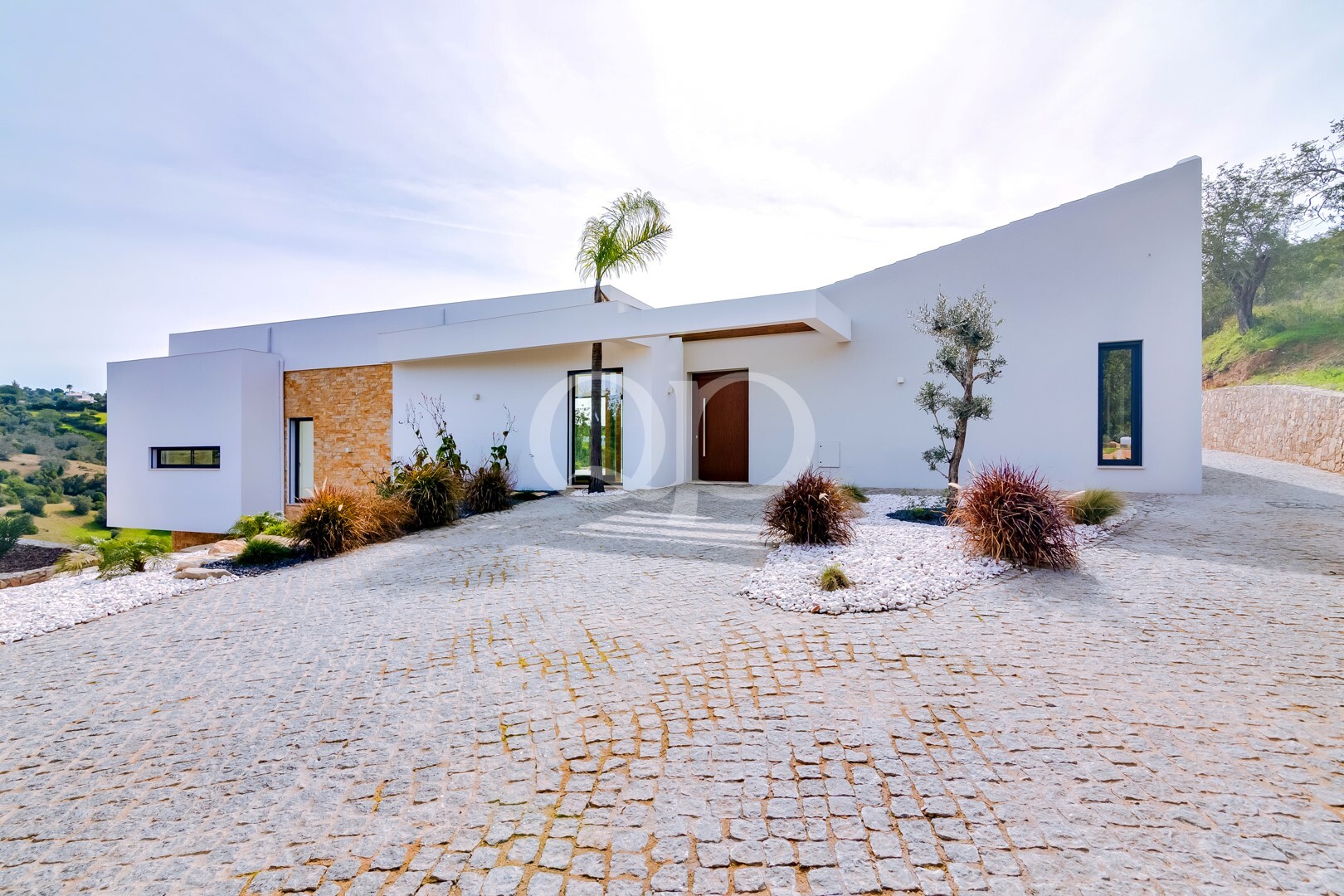 Modern white, single-story home with a stone accent wall, palm tree, and gravel landscaping on a cobblestone driveway.