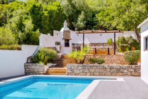 Outdoor pool area with white walls, stone accents, and a built-in grill, surrounded by lush greenery and steps leading to a patio.