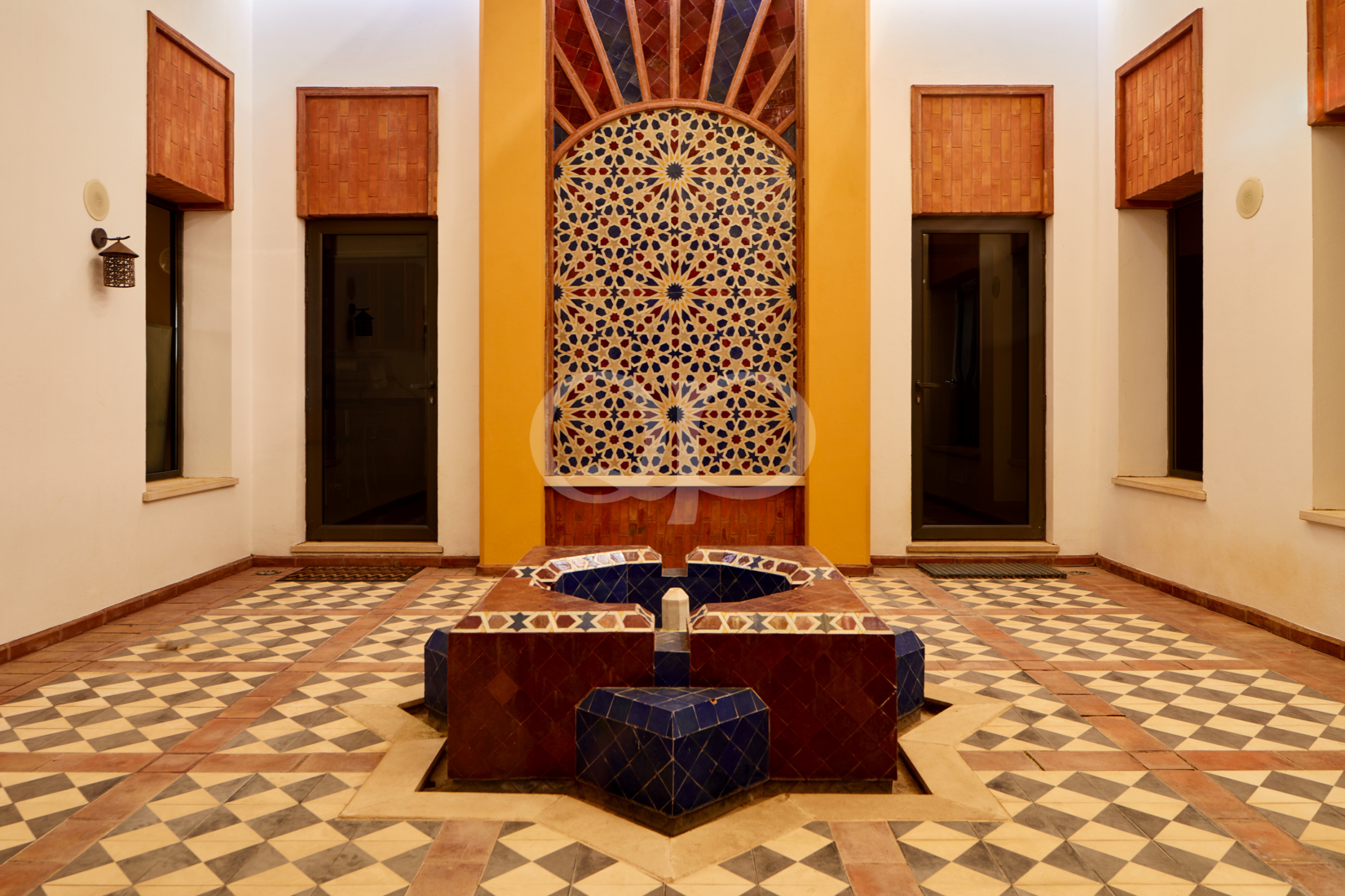 Pátio interior com uma fonte geométrica no centro, revestida de azulejos azuis, e uma parede de mosaico colorido emoldurada por pilares laranja de cada lado.
