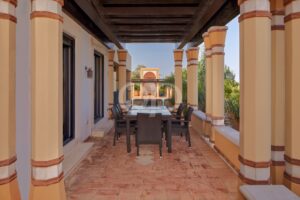 Terraço exterior com colunas em bege e terracota e uma pérgula em madeira escura, com uma mesa de jantar e cadeiras em vime preto ao longo de um corredor.