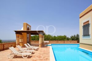 Área junto à piscina com uma piscina azul, arcos laranja e espreguiçadeiras brancas sob um céu límpido