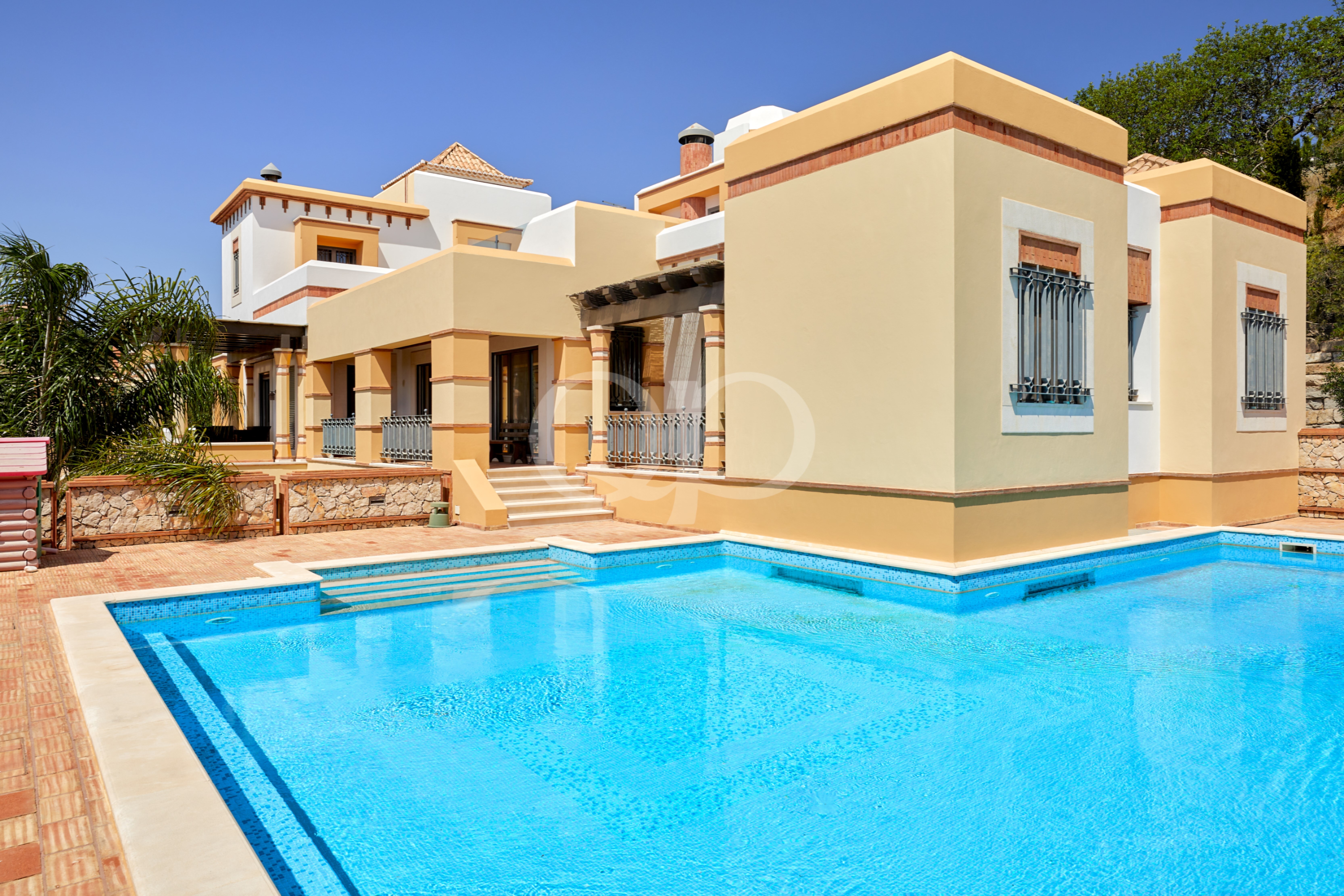 Villa de villégiature avec, au premier plan, une piscine bleue et des bâtiments de couleur jaune crème sous un ciel bleu limpide
