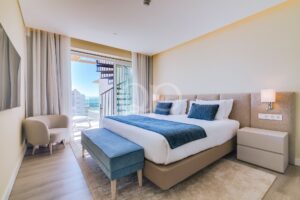 Ein Hotelzimmer mit einem großen Bett, blauen Akzenten und einer Balkontür, die einen sonnigen Blick auf die umliegenden Gebäude und das Meer bietet.