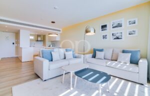 Ein offen gestaltetes Wohnzimmer mit einem beigen Ecksofa, blau-weißen Kissen und zwei Couchtischen.
