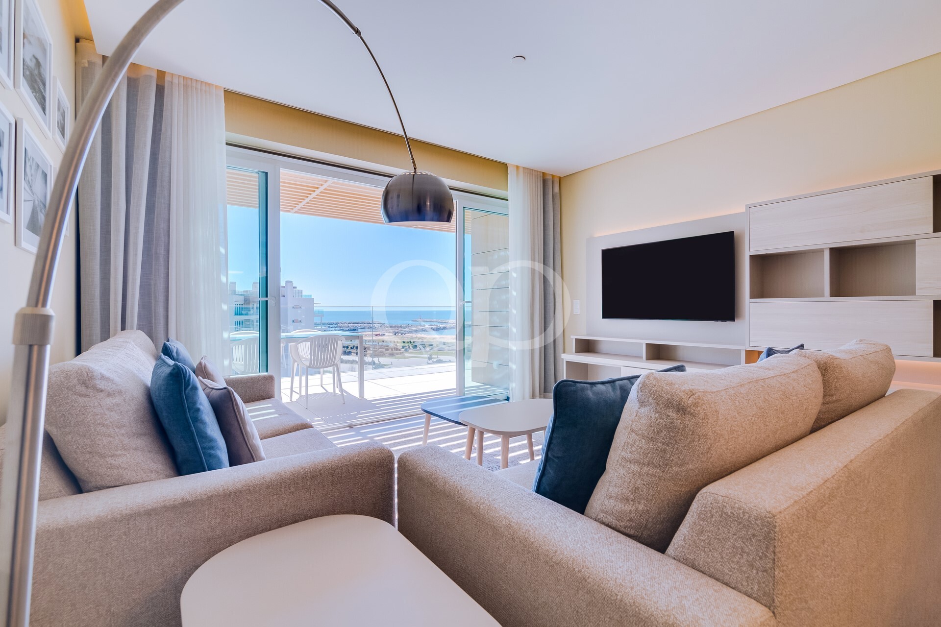 Sala de estar acolhedora em tons de bege, com almofadas azuis, em frente a uma televisão fixada na parede e a um móvel de arrumação em madeira clara; as portas da varanda abrem-se para uma vista para o mar.
