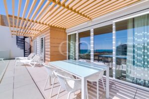 Moderne Außenterrasse mit weißen Liegestühlen und einem gläsernen Esstisch unter einer Pergola aus Holzlatten mit Blick auf das Meer.