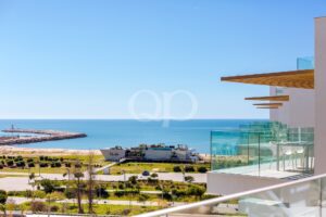 Modernes Gebäude mit Glasfassade und orangefarbenen, horizontalen Vordächern entlang eines Balkons mit Blick auf das blaue Meer und die Mole.