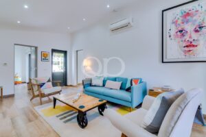 Bright living room with light wood floor, blue sofa, beige armchairs, colorful rug, and abstract portrait on wall.