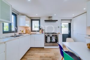 Bright, modern white kitchen with a stainless steel oven, white cabinets, and a central island; colorful chairs around a white dining table nearby.