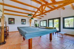 Bright game room with a blue-covered pool table under exposed wooden ceiling beams and green pendant lights.
