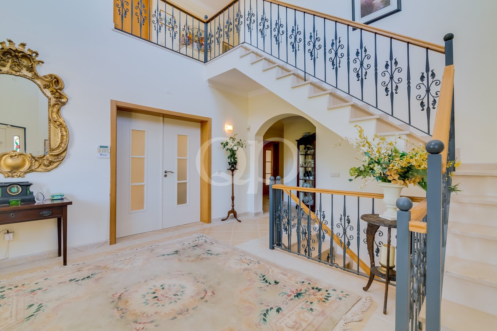 Um hall de entrada luminoso de dois andares, com portas duplas de painéis de vidro, um espelho ornamentado dourado e uma mesa consola de madeira junto à entrada.