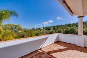 Varanda com ladrilhos de terracota, paredes brancas e uma coluna, com vista para uma encosta verdejante sob um céu azul brilhante.