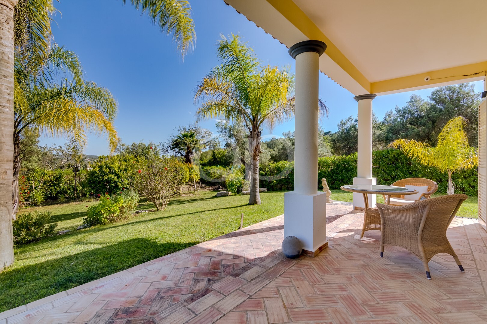 Pátio com cadeiras de vime e uma mesa redonda de vidro sob uma varanda coberta de lona amarela, com vista para um jardim exuberante com palmeiras e um céu azul brilhante.
