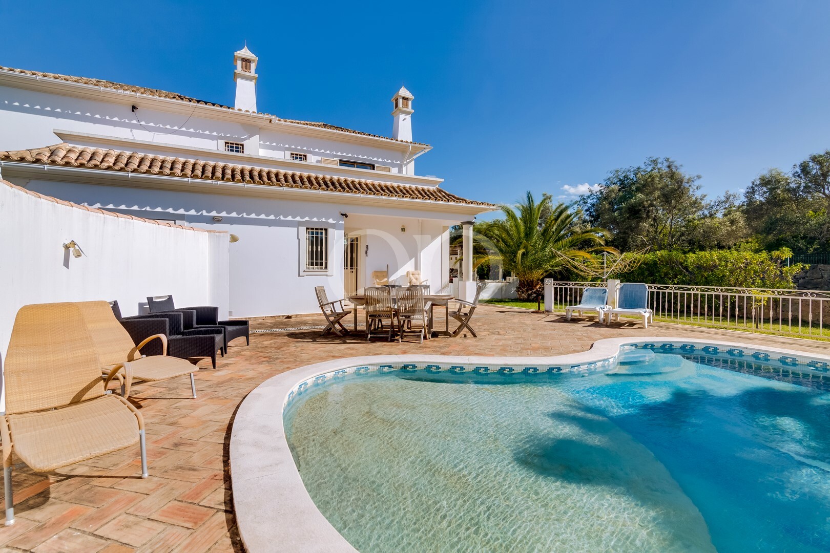 Moradia branca com telhado de terracota, mesa de jantar ao ar livre e piscina sob um céu azul límpido. Um cenário relaxante no pátio, com espreguiçadeiras e palmeiras.