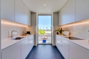 Contemporary white galley kitchen with sleek cabinets and under-cabinet lighting, opening to a sunny balcony with ocean view.