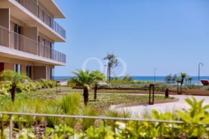 Apartment building with stacked balconies along a landscaped garden, ocean visible in the distance on a sunny day.
